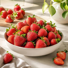 A white ceramic bowl filled with fresh red strawberries, with a few whole and sliced strawberries placed on a light marble surface. A wooden board with more strawberries and a potted plant are softly blurred in the background.