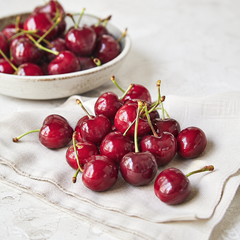 Red cherries on a white cloth with a bowl of cherries in the background