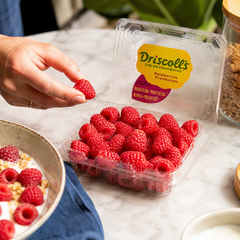 Driscoll's raspberries in a clear plastic container on a marble surface with a hand picking a raspberry.